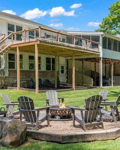 Modern farmhouse with stone foundation, upper deck, turquoise Adirondack chairs on lower patio, sunset sky, manicured lawn
