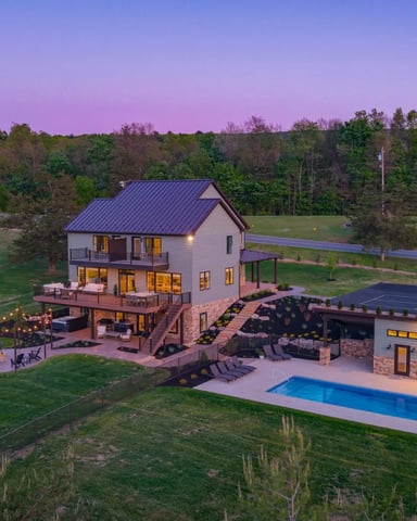 Modern farmhouse with metal roof on expansive grounds at dusk, featuring illuminated windows, pool, patio with string lights, and surrounding forest.