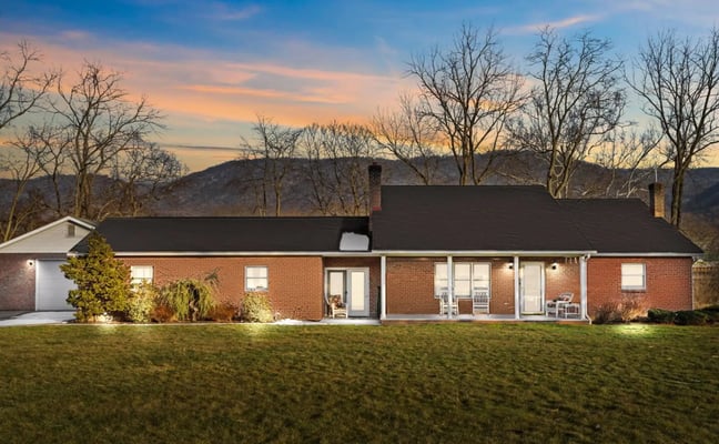 Log cabin with red metal roof and wraparound deck at dusk, surrounded by trees and pastoral landscape